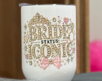 May include: A white tumbler featuring the text BRIDE STATUS: ICONIC in gold lettering. The design is embellished with rhinestones, a gold crown, a pink satin bow, and small pearl accents. A clear plastic lid and a clear straw are included with the cup.