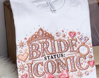 May include: A white t-shirt featuring a graphic design with the text "BRIDE STATUS: ICONIC" in rose gold, jewel-encrusted lettering. The design includes a tiara, a diamond ring, pink hearts, pearls, and a pink satin bow at the bottom center.