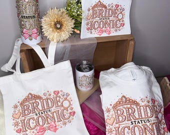 May include: A white tote bag, a t-shirt, a tall tumbler, a small tumbler, and a zippered pouch, all featuring a graphic of a tiara, a diamond ring, hearts, and a pink bow with the text BRIDE STATUS: ICONIC in rose gold and pink tones.