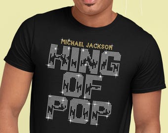 以下が含まれることがあります： 黒いTシャツ。テキスト「MICHAEL JACKSON KING OF POP」がキラキラ光るラインストーンのようなデザインで施されています。テキストは太字で、金色のアクセントが付いています。Tシャツは、クラシックなクルーネックです。