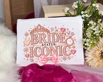 May include: A white rectangular zippered pouch featuring a rose gold and pink graphic design with the text "BRIDE STATUS: ICONIC". The design includes a tiara, a diamond ring, hearts, and stars, accented with a pink satin bow at the bottom center.