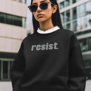 May include: A black sweatshirt with the word "resist" spelled out in rhinestones. The person in the image is wearing sunglasses and a white collared shirt underneath the sweatshirt. The background is a blurred city scene.