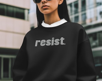 以下が含まれることがあります： ラインストーンで「resist」と書かれた黒いスウェットシャツ。画像内の人物はサングラスと白い襟付きシャツをスウェットシャツの下に着用しています。背景はぼやけた都市のシーンです。