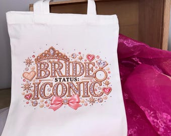 May include: A white canvas tote bag featuring a graphic print with the text "BRIDE STATUS: ICONIC". The design includes a gold crown, a diamond ring, pink hearts, sparkling star accents, and a pink bow, all rendered in a jeweled aesthetic.