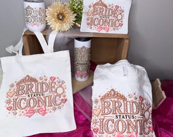 May include: A white tote bag, a white t-shirt, a zippered pouch, and two tumblers, all featuring a rose gold and pink graphic design with the text "BRIDE STATUS: ICONIC," a tiara, a diamond ring, and a pink bow, arranged with a gold flower.