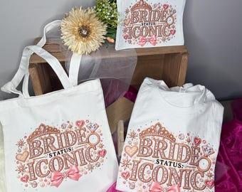 May include: A white canvas tote bag, a white zippered pouch, and a white t-shirt, each featuring a graphic print of a rose gold tiara, a diamond ring, hearts, pearls, and a pink bow, with the text "BRIDE STATUS: ICONIC" in rose gold lettering.