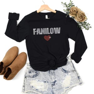 K&ouml;nnte beinhalten: Schwarzes Langarmshirt mit dem Wort "FANILOW" in Strassbuchstaben und einem roten Strassherz. Das Shirt wird mit Jeansshorts und braunen Stiefeletten kombiniert. Das Shirt h&auml;ngt an einem Holzkleiderb&uuml;gel.