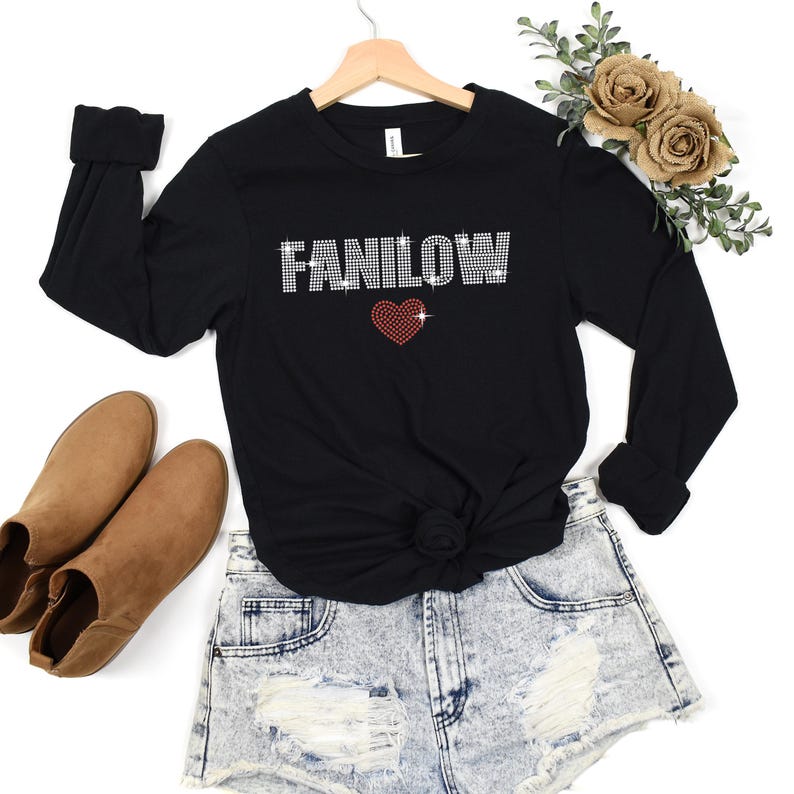 K&ouml;nnte beinhalten: Schwarzes Langarmshirt mit dem Wort "FANILOW" in Strassbuchstaben und einem roten Strassherz. Das Shirt wird mit Jeansshorts und braunen Stiefeletten kombiniert. Das Shirt h&auml;ngt an einem Holzkleiderb&uuml;gel.