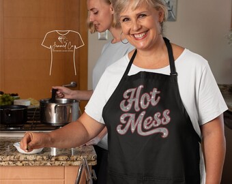 以下が含まれることがあります： キラキラ光る筆記体フォントで「Hot Mess」と書かれた黒いエプロン。キッチンで人が着用しています。背景には別の人が見えます。エプロンには首ひもとフロントポケットが付いています。