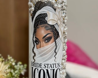 May include: A white cylindrical tumbler featuring a graphic illustration of a person with braided hair wearing a veil and a bow. The design includes the text "BRIDE STATUS: ICONIC" above a gold ring icon. The top is adorned with white pearls and clear rhinestones.