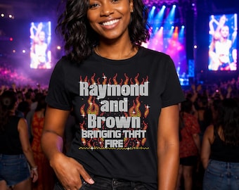以下が含まれることがあります： ラインストーンのデザインで「Raymond and Brown Bringing That Fire」という文字が入った黒いTシャツ。テキストは赤、黄、オレンジ色のラインストーンの炎で囲まれています。Tシャツは群衆の前で人が着ています。