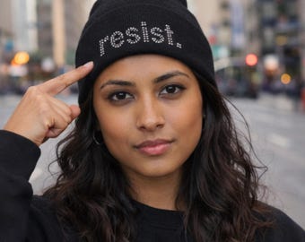 以下が含まれることがあります： ラインストーンで「resist」と書かれた黒いビーニー帽。ぼやけた街並みを背景に、人がかぶっています。その人はビーニー帽を指さしています。