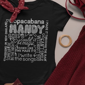 May include: Black t-shirt with rhinestone text that reads "Copacabana Mandy Shoops Could It Be Magic Looks Like We Made It I Write The Songs." The shirt has a crew neck and short sleeves. The text is in a variety of fonts and sizes.