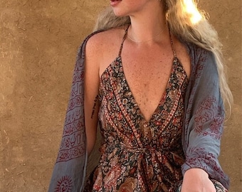 May include: A woman wearing a long, flowing, dark floral print dress with a grey shawl. She has long blonde hair with braids and is wearing several rings on her fingers.