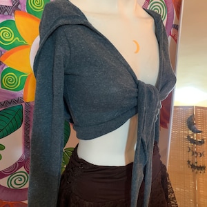 Könnte beinhalten: Ein dunkelgraues, langärmliges Crop-Top mit Bindeband vorne und Kapuze. Das Oberteil ist aus weichem, dehnbarem Stoff und hat eine lockere Passform.