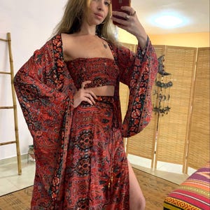 May include: A three-piece bohemian outfit in shades of red, black, and teal. The set includes a crop top, a long skirt with a thigh-high slit, and a kimono-style jacket with wide sleeves. The fabric features an intricate paisley pattern.