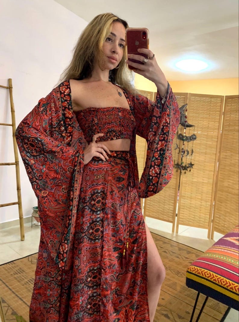 May include: A three-piece bohemian outfit in shades of red, black, and teal. The set includes a crop top, a long skirt with a thigh-high slit, and a kimono-style jacket with wide sleeves. The fabric features an intricate paisley pattern.