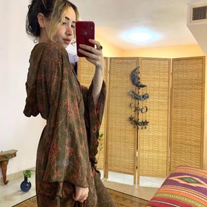 May include: A long, flowing brown robe with a floral pattern in red. The robe has wide sleeves and is worn by a person taking a mirror selfie. The background includes a wooden screen and a colorful patterned cushion.