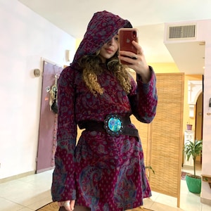 Puede incluir: Un vestido morado con capucha con estampado paisley y un cinturón negro con hebilla turquesa y plateada. El vestido tiene mangas largas y un corte holgado.