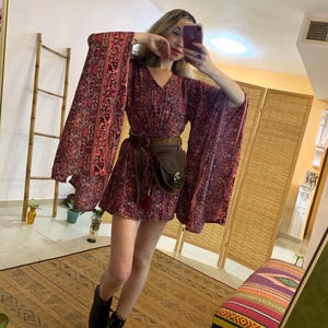 Puede incluir: Un minivestido burdeos con estampado floral y mangas anchas. El vestido está ceñido a la cintura con un cinturón marrón y una riñonera a juego. Las botas de combate negras completan el look bohemio.
