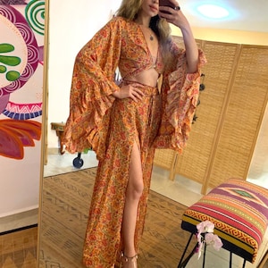 May include: An orange floral print two-piece set with a crop top and a long skirt. The top has long, flared sleeves and a tie front. The skirt has a high slit. The outfit is made of a lightweight fabric.