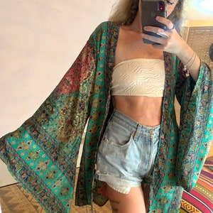 Op de afbeelding: Een tealblauwe en gouden kimono met lange mouwen en een wijd, vloeiende silhouet. De kimono wordt gedragen over een witte bandeau top en denim shorts.