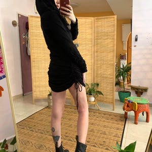 May include: Black hooded dress with long sleeves and a ruched skirt. The dress features a drawstring detail at the hem and is paired with black combat boots. The outfit is captured in a full-length mirror shot.
