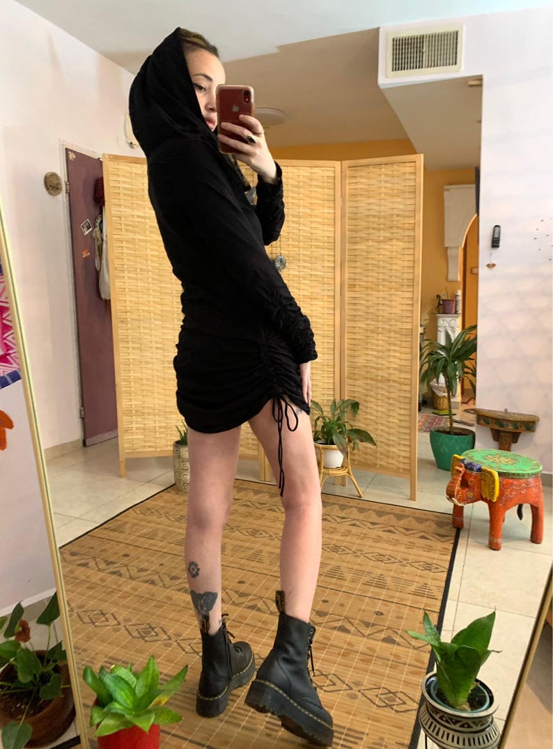 May include: Black hooded dress with long sleeves and a ruched skirt. The dress features a drawstring detail at the hem and is paired with black combat boots. The outfit is captured in a full-length mirror shot.