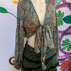 May include: A paisley print top with long sleeves and a tie front, paired with a green skirt featuring black stripes and a tassel. The top has a V-neck and a mix of blue, brown, and red colours.