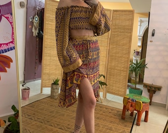 Könnte beinhalten: Ein zweiteiliges Boho-Outfit mit schulterfreiem Crop-Top und passendem Rock. Das Set ist mit gelben, lila und roten Mustern versehen. Der Rock hat einen hohen Schlitz. Das Outfit wird mit High-Heel-Sandalen getragen.