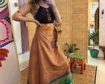 May include: A woman wearing a black crop top and a long, patterned wrap skirt. The skirt is primarily orange with a geometric design and features green and blue stripes at the hem. A yellow sash is tied around the waist.