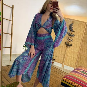 May include: A two-piece set featuring a crop top and wide-leg pants. The set is made of a flowing fabric with a swirling pattern in shades of blue, purple, and teal. The top has long, flared sleeves and a knotted front. The pants are cropped at the calf.