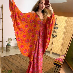 Może przedstawiać: Długa, zwiewna sukienka maxi w pomarańczowo-różowy wzór kwiatowy z głębokim dekoltem w kształcie litery V i rozkloszowanymi rękawami. Sukienka wykonana jest z lekkiej tkaniny i ma luźny krój.