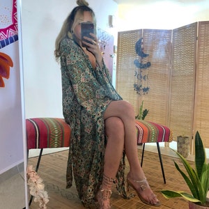 May include: A woman wearing a long, flowing green and floral patterned robe with a plunging neckline. She is sitting on a colorful striped bench with her legs crossed. She is wearing clear heeled sandals with ankle straps.