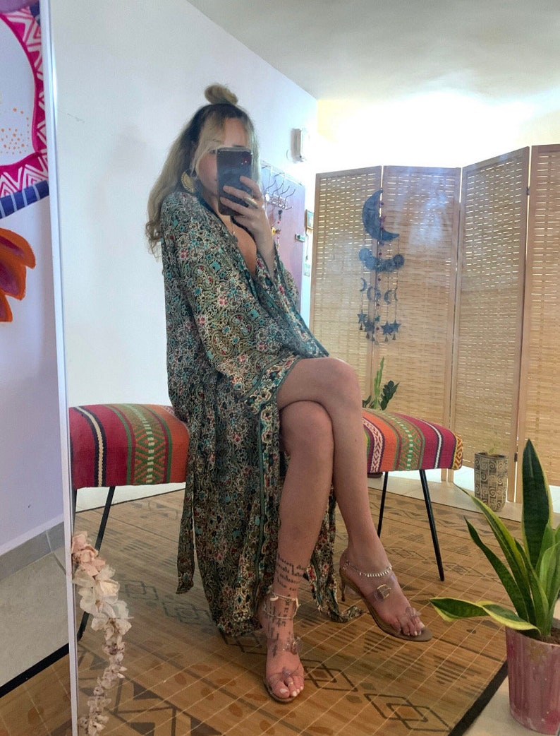 May include: A woman wearing a long, flowing green and floral patterned robe with a plunging neckline. She is sitting on a colorful striped bench with her legs crossed. She is wearing clear heeled sandals with ankle straps.