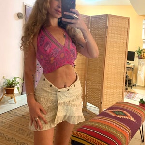 Puede incluir: Una mujer lleva un top halter estampado en rosa y verde con una falda de crochet blanca. La falda tiene un dobladillo con volantes y un cierre de botón. La mujer está de pie frente a una pantalla de madera.
