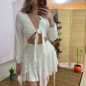 Puede incluir: Conjunto de dos piezas color crema. La parte superior es un top corto de manga larga con lazo. La falda es una minifalda con capas, detalles de encaje y lazos laterales. El conjunto está hecho de una tela suave y fluida, ideal para un look bohemio.