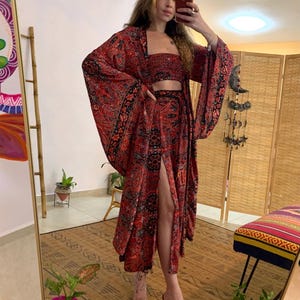 May include: A three-piece bohemian outfit in red with a black paisley pattern. The set includes a crop top, a long skirt with a high slit, and a kimono-style jacket with wide sleeves. The outfit is worn with clear heels.