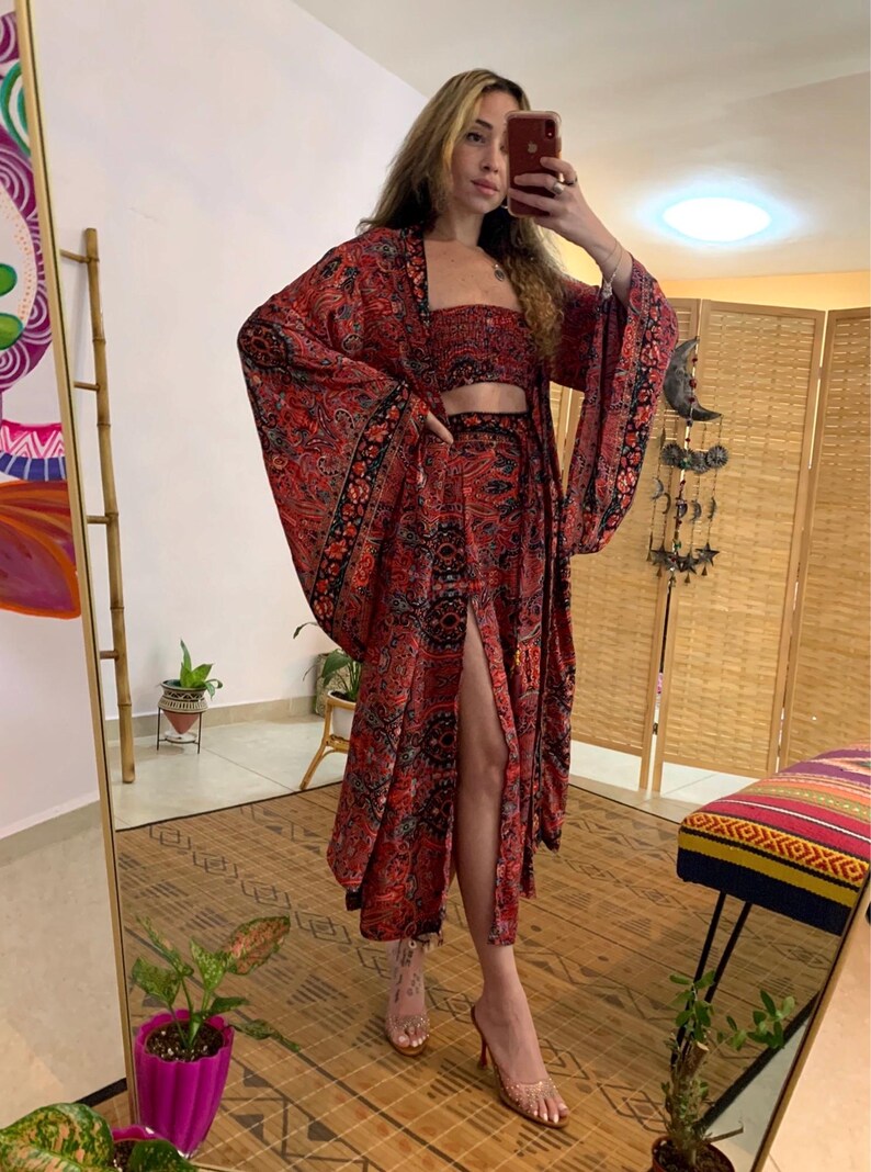 May include: A three-piece bohemian outfit in red with a black paisley pattern. The set includes a crop top, a long skirt with a high slit, and a kimono-style jacket with wide sleeves. The outfit is worn with clear heels.