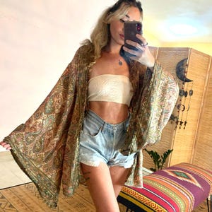 May include: A sheer, open-front kimono in shades of brown, green, and gold with a paisley pattern. The kimono has wide sleeves and is worn over a white tube top and denim shorts. The outfit is completed with a bench with a colorful, patterned cushion.