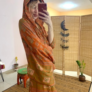May include: An orange and green patterned hooded garment. The fabric features a repeating design of floral and geometric shapes. The garment appears to be made of a lightweight material, suitable for layering or warmer weather.
