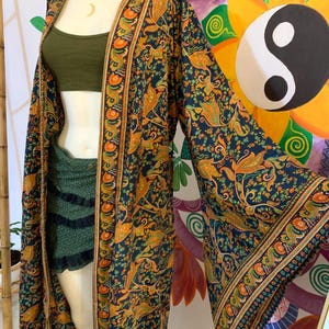 Può includere: Una lunga vestaglia stile kimono con sfondo blu scuro e un intricato motivo paisley nelle tonalità dell'arancione e dell'oro. La vestaglia ha maniche ampie e un bordo decorativo. L'indumento è aperto sul davanti, rivelando un top e pantaloncini verdi.