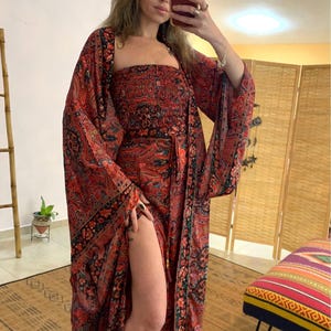 May include: A red paisley print ensemble featuring a strapless top, a long skirt with a high slit, and a matching kimono-style jacket. The outfit has black and green accents, creating a bohemian aesthetic. The dress is floor length.