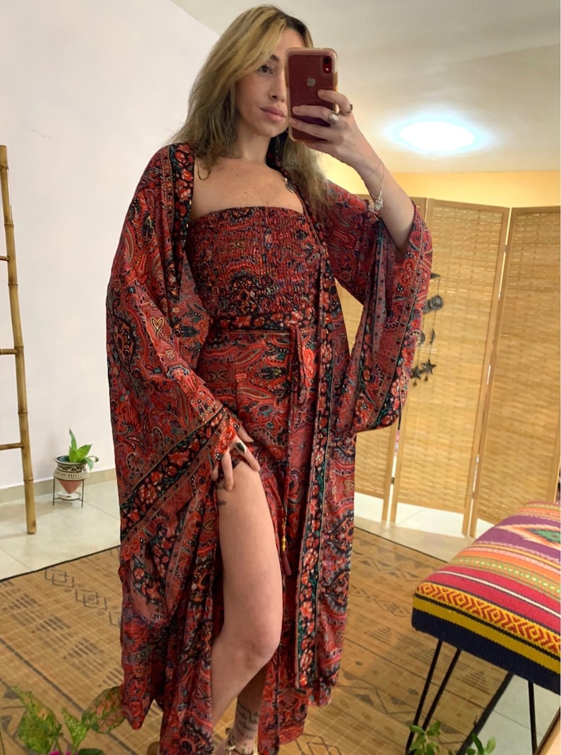 May include: A red paisley print ensemble featuring a strapless top, a long skirt with a high slit, and a matching kimono-style jacket. The outfit has black and green accents, creating a bohemian aesthetic. The dress is floor length.