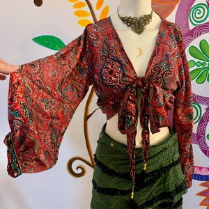 Könnte beinhalten: Ein rotes Oberteil mit Paisley-Muster, langen, ausgestellten Ärmeln und einem Bindeband vorne. Das Oberteil wird mit einem grünen Rüschen-Minirock kombiniert. Eine dekorative Halskette befindet sich um den Hals der Schaufensterpuppe.