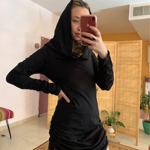 May include: A black hooded dress with long sleeves and a ruched skirt. The dress has a drawstring detail on the side and is made of a soft, flowing fabric. The hood is draped over the head.