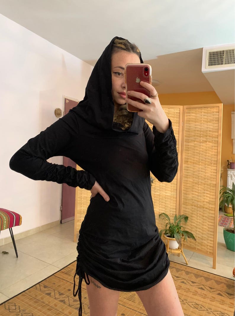 May include: A black hooded dress with long sleeves and a ruched skirt. The dress has a drawstring detail on the side and is made of a soft, flowing fabric. The hood is draped over the head.