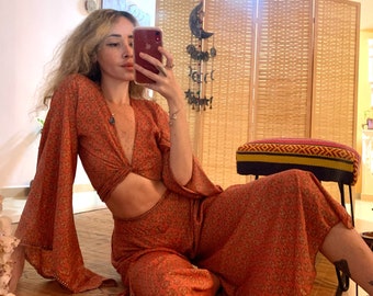 May include: A woman wearing a two-piece outfit with a floral print. The top is a crop top with a twist detail in the front. The pants are wide-legged and have a flowy silhouette. The outfit is in shades of orange and brown.