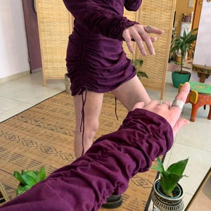 May include: A plum-colored, long-sleeved dress with ruched detailing on the sleeves and sides. The dress has a drawstring at the hem, creating a gathered effect. The fabric appears to be a soft, jersey-like material. The dress is shown in a room with a natural wood screen.