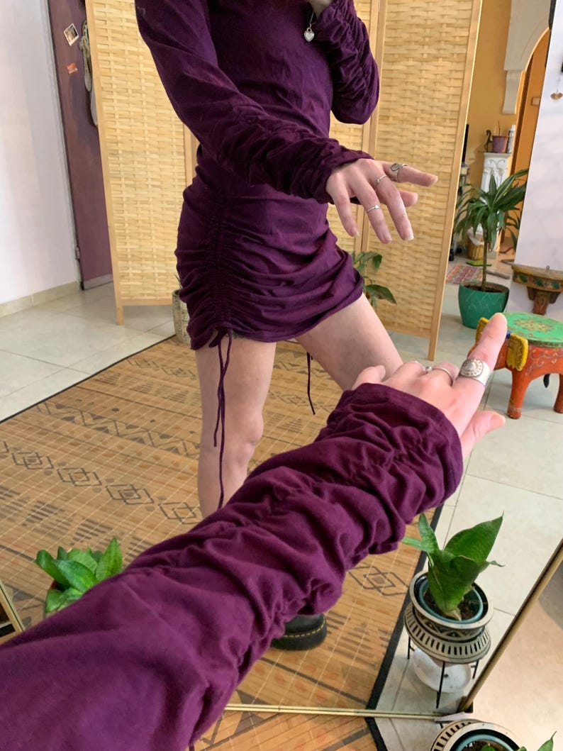 May include: A plum-colored, long-sleeved dress with ruched detailing on the sleeves and sides. The dress has a drawstring at the hem, creating a gathered effect. The fabric appears to be a soft, jersey-like material. The dress is shown in a room with a natural wood screen.